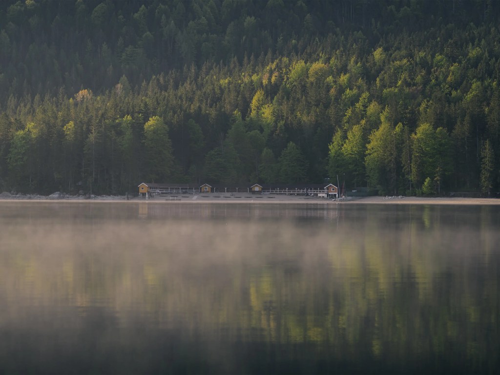 Blick über den Eibsee zu einem Bootshaus mit Spiegelung der Wälder.