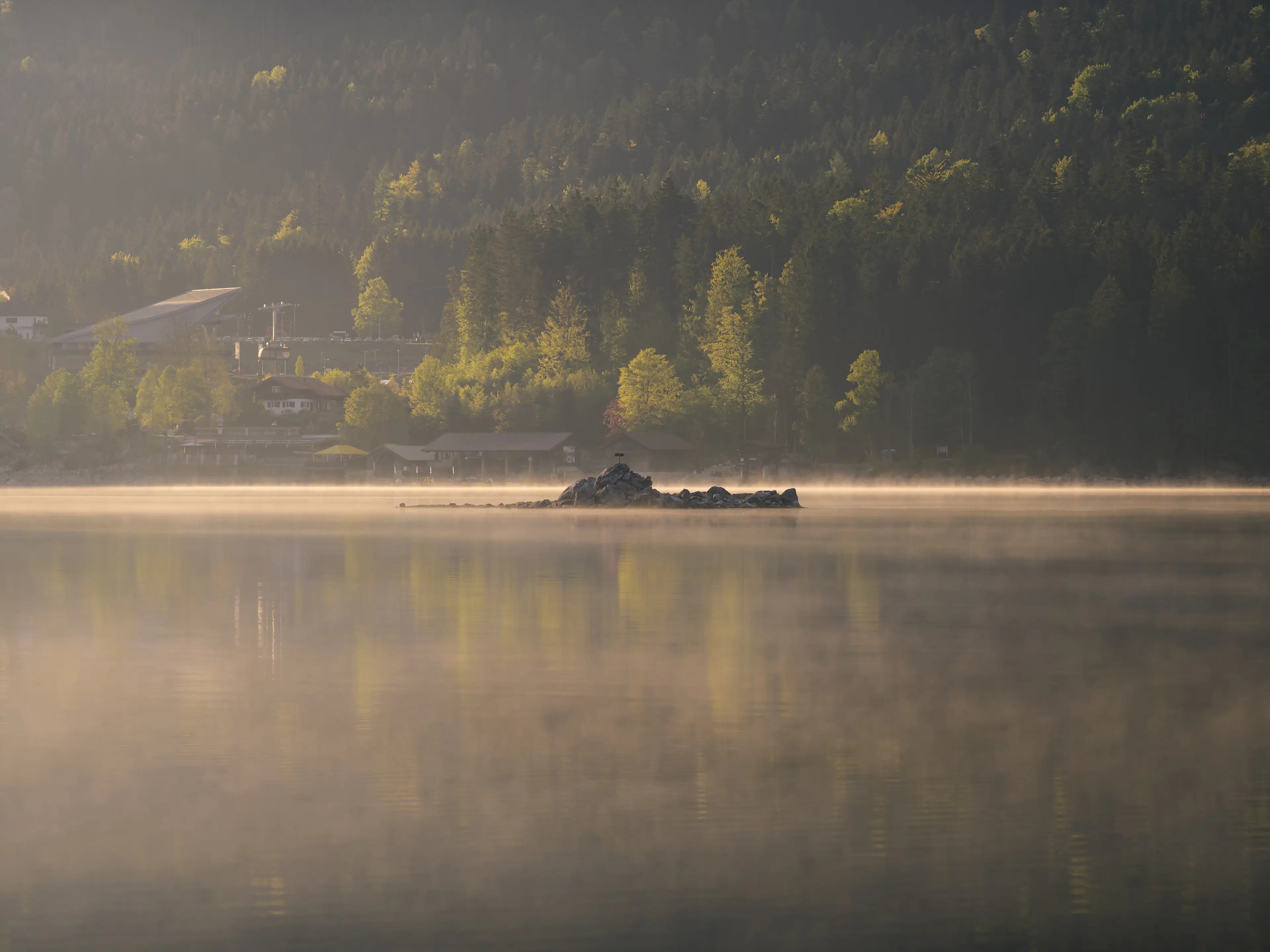 Eine kleine Insel aus Steinen ragt aus dem Wasser, über dem leichter Morgennebel liegt.