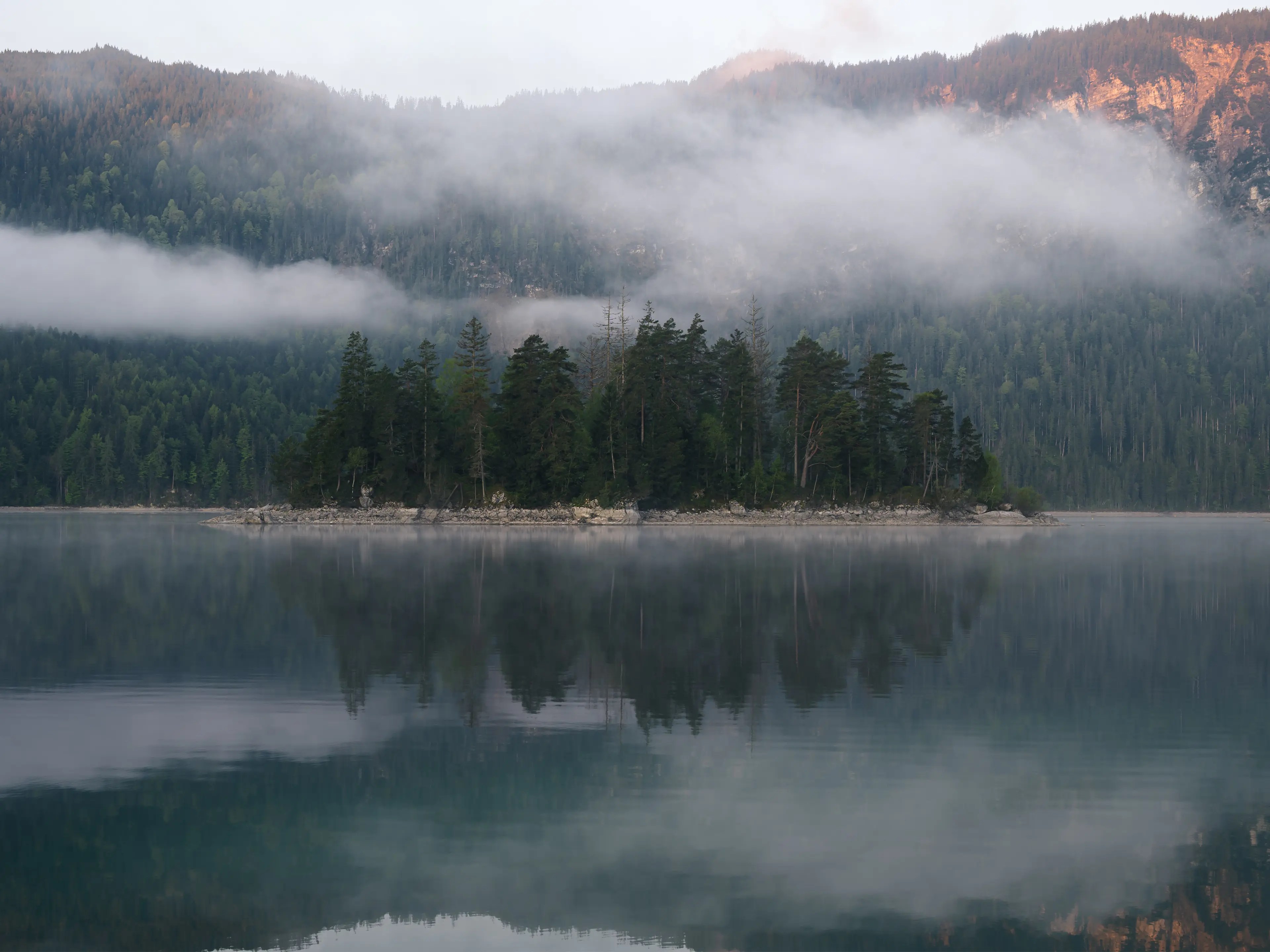 Blick zur Sasseninsel im Eibsee, die sich im See spiegelt. Über der Insel liegt leichter Nebel.