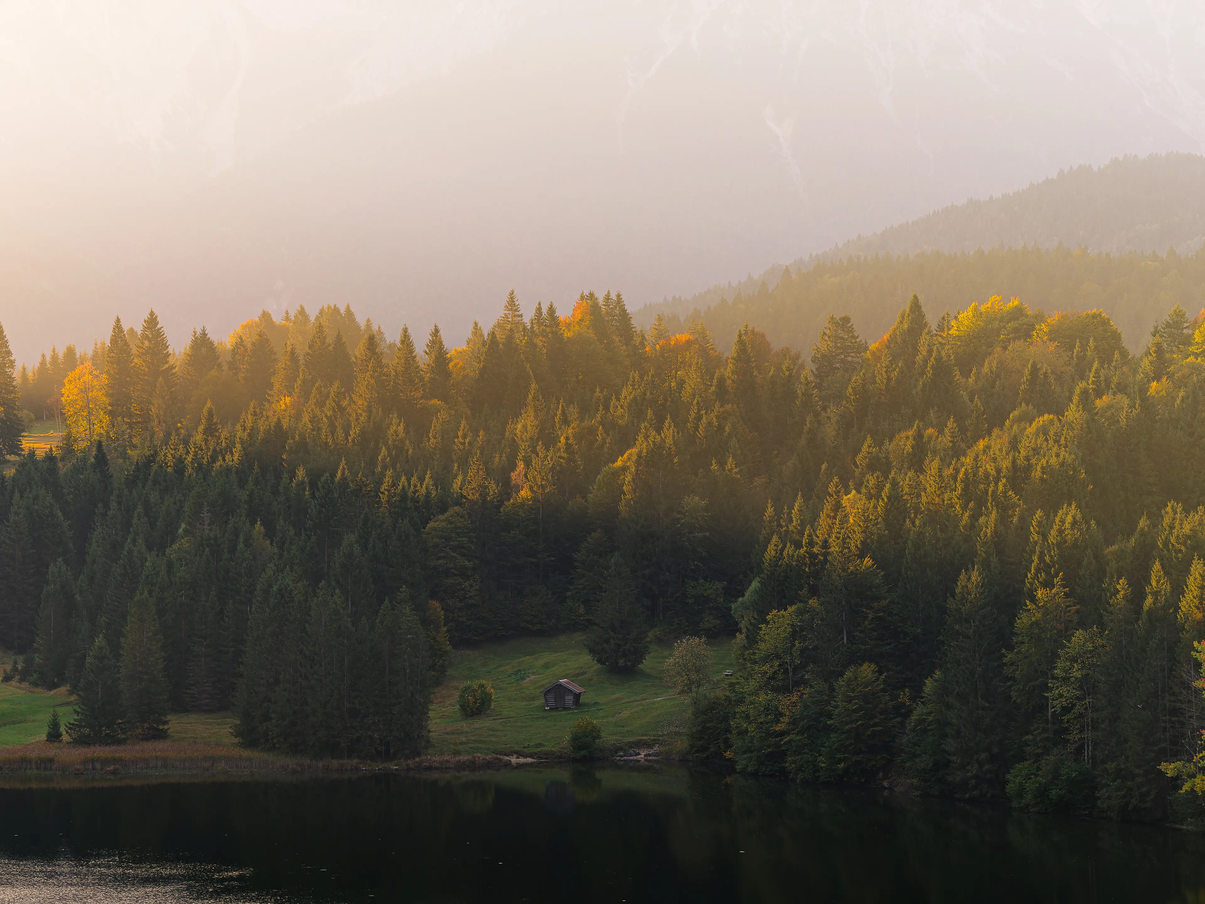 Warmes Licht eines herbstlichen Sonnenaufgangs erleuchtet einen kleinen Wald an einem See, an dem eine kleine Hütte zu erkennen ist.
