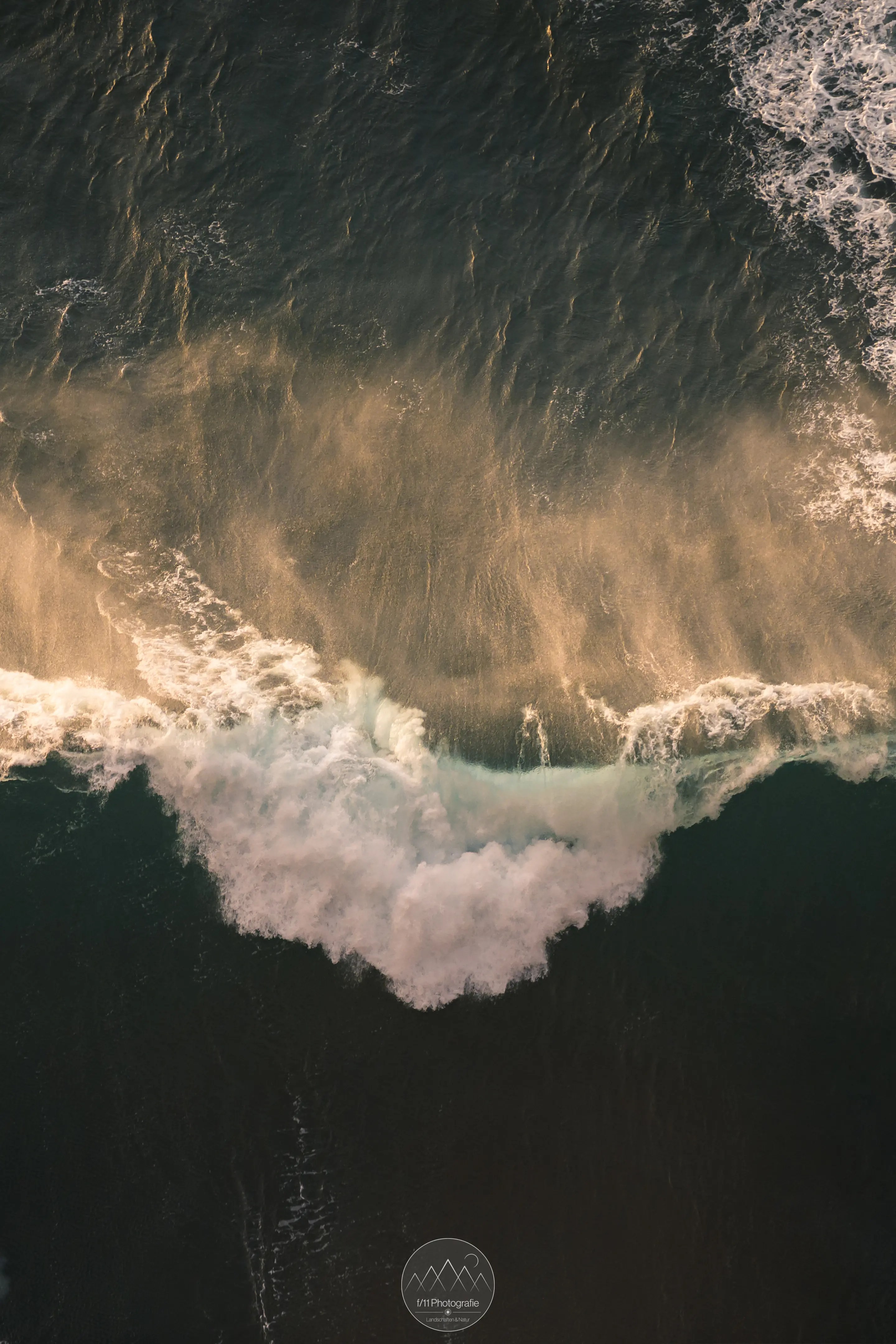 Eine Welle bricht an der Küste, fotografiert direkt von oben herab.