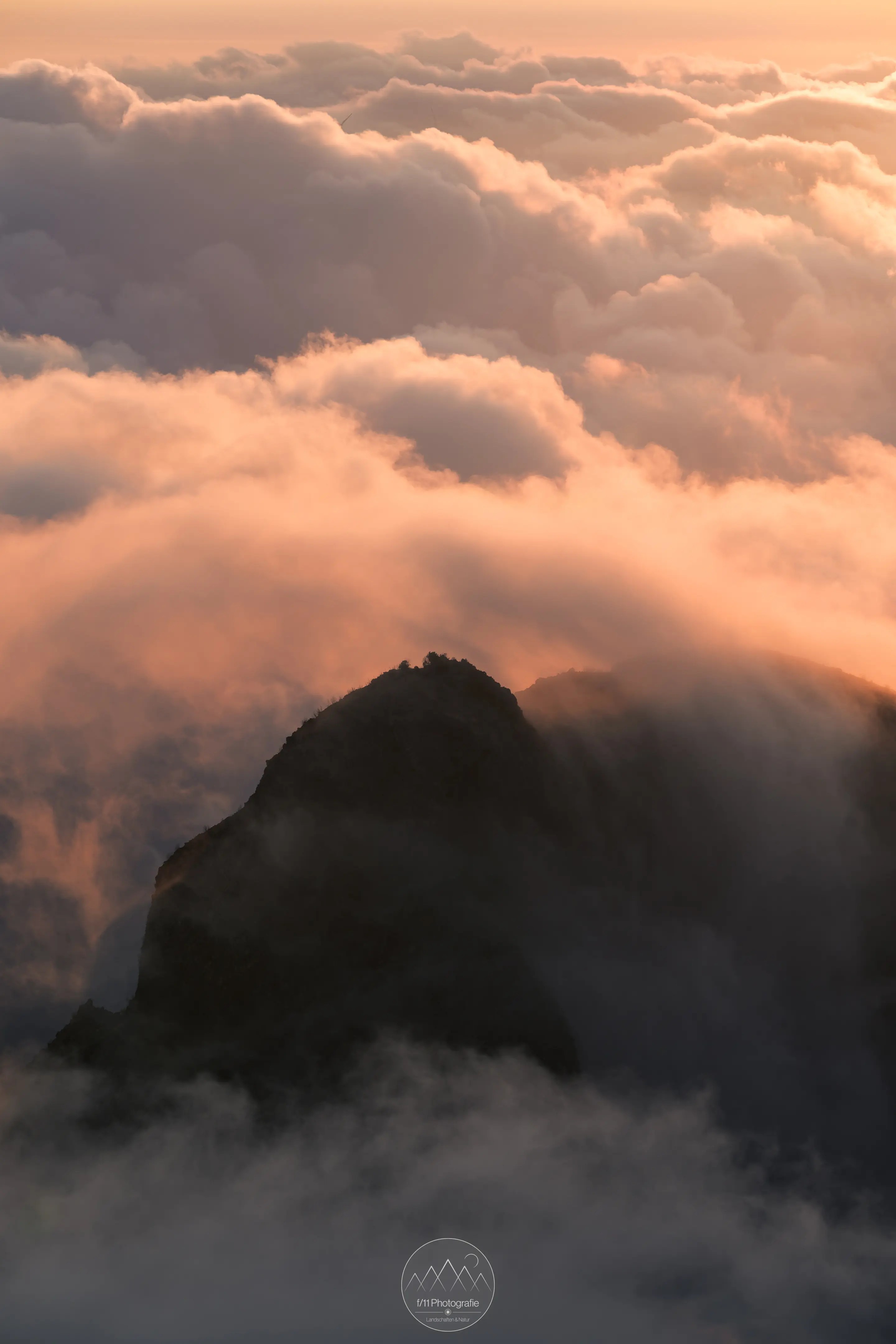Berggipfel umgeben von Wolken, die orange im Licht der untergehenden Sonne leuchten.