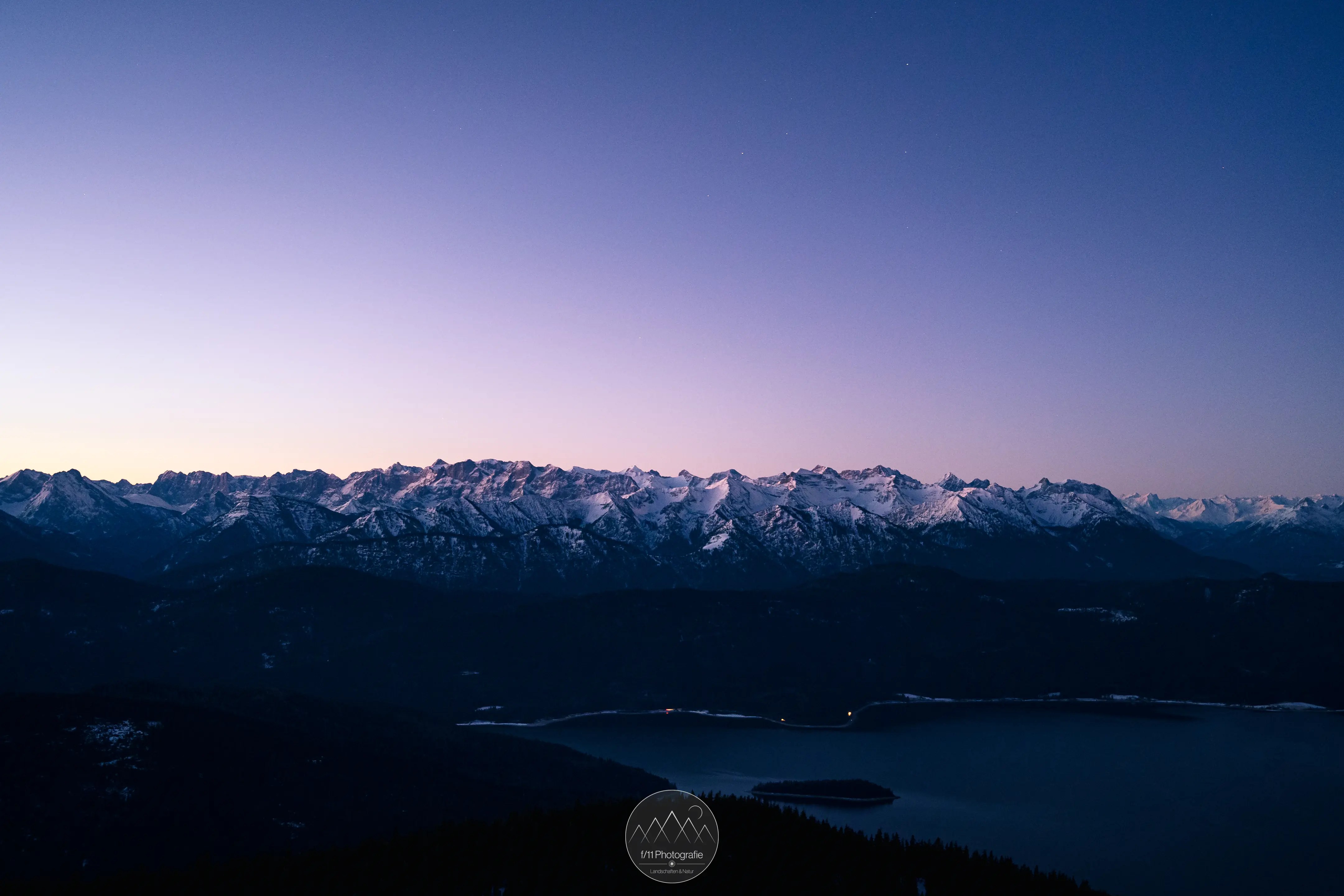 Blick auf die verschneiten Berggipfel des Karwendelgebirges zur blauen Stunde. Im Vordergrund ist der Walchensee zu sehen. 